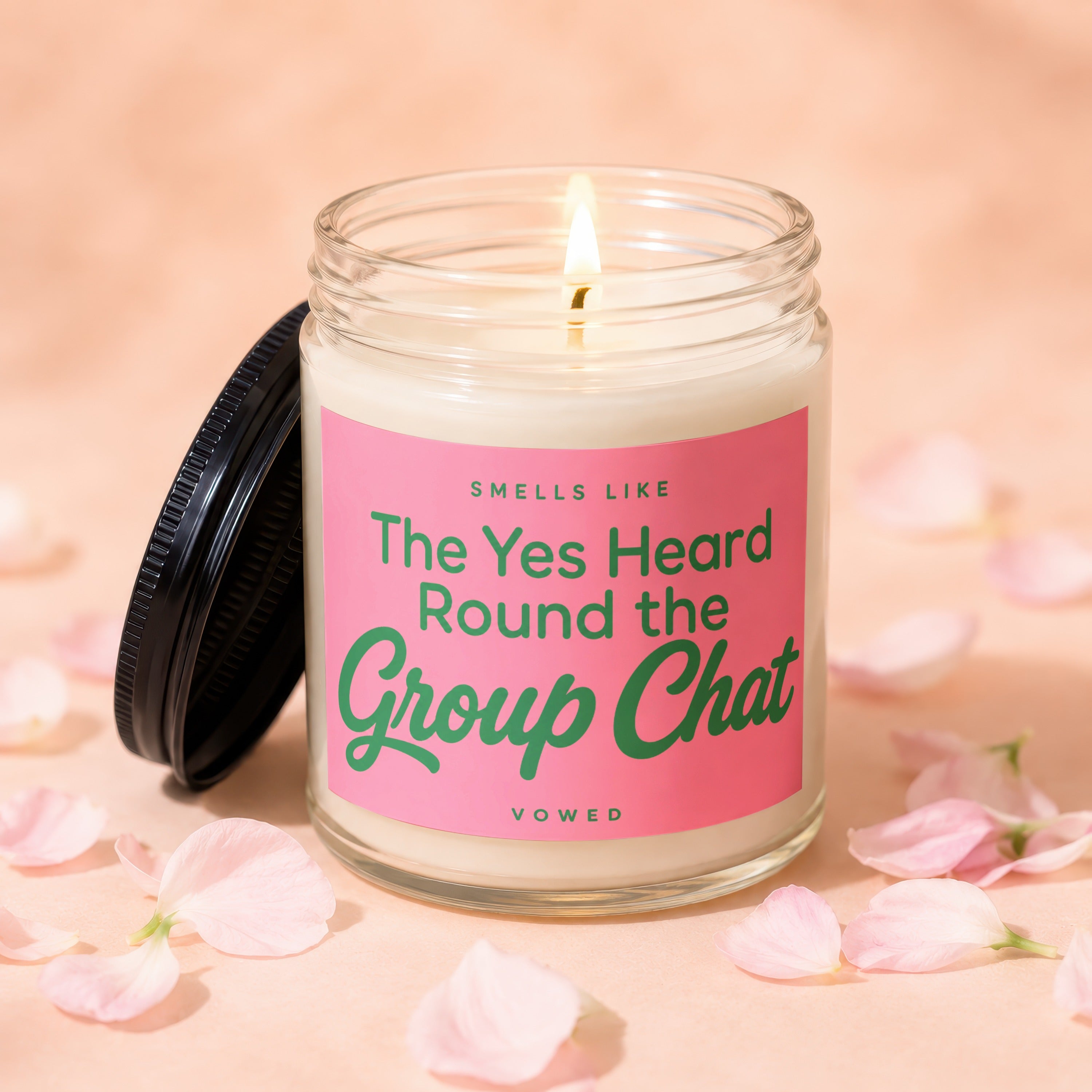 Glass candle with lit wick and pink label that reads "Smells Like The Yes Heard Round the Group Chat".