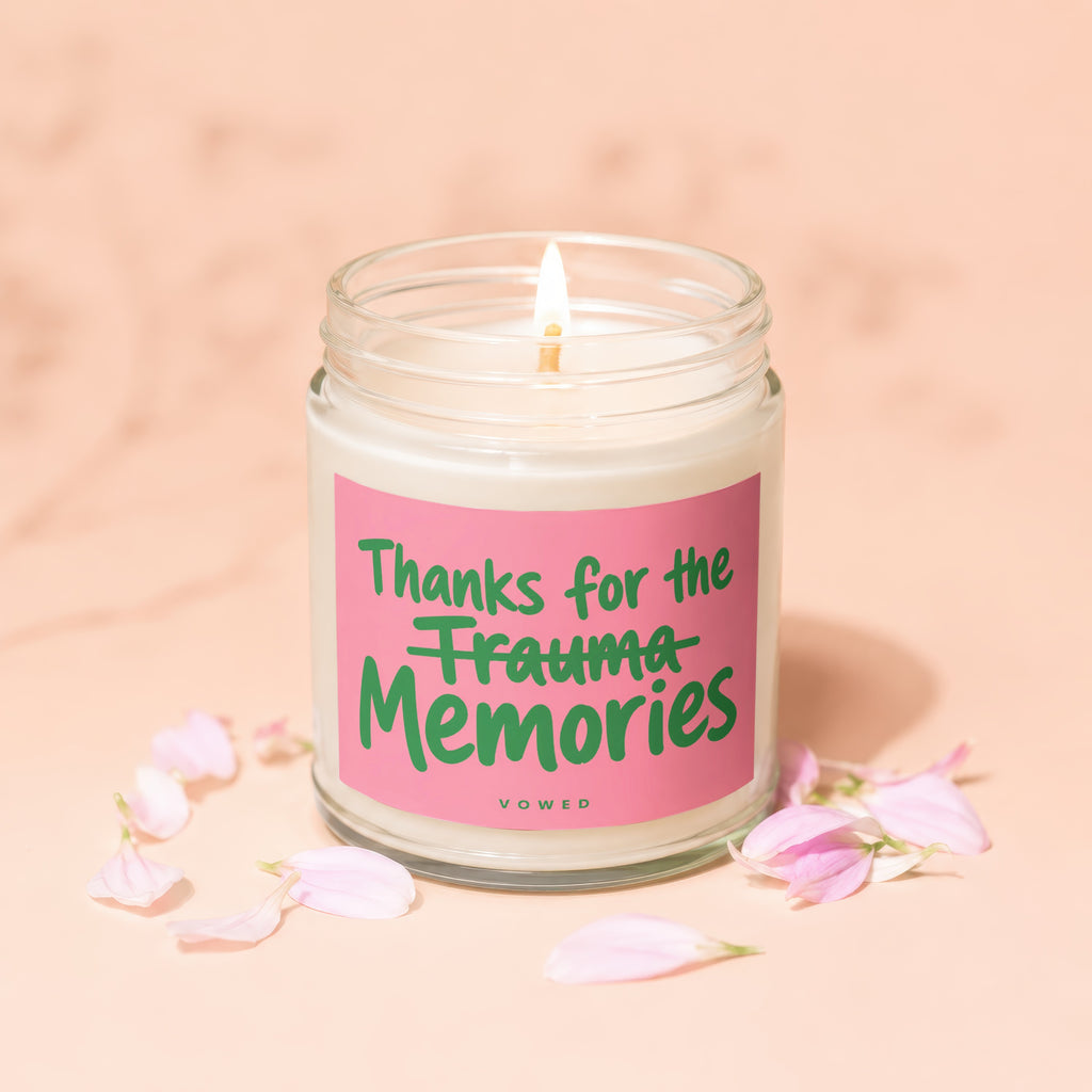 A lit candle with pink label reading "Thanks for the Trauma Memories," surrounded by pink petals.