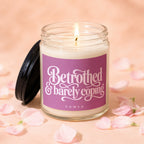 Glass candle with lit wick on peach background with pink petals, reads "Betrothed & barely coping VOWED".