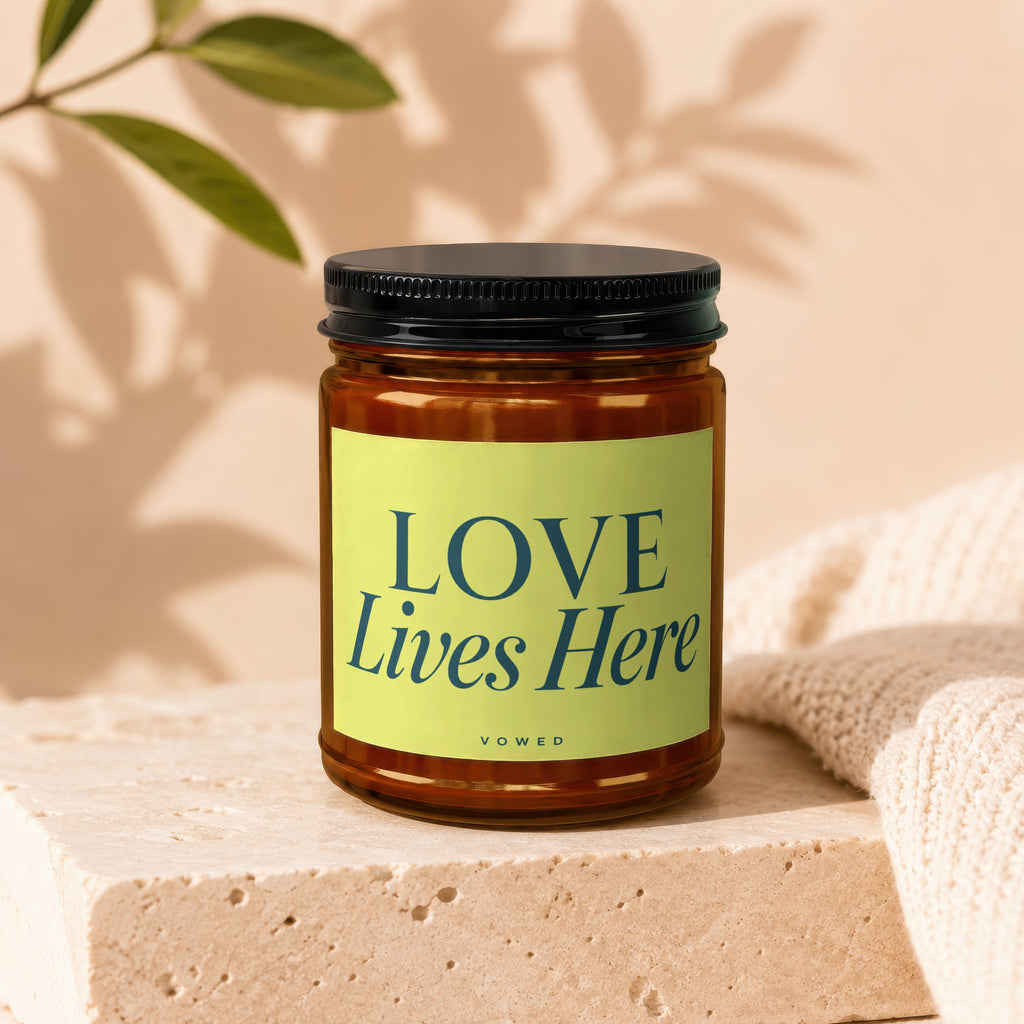 Amber candle in jar with "LOVE Lives Here" label, on cream stone and textile.