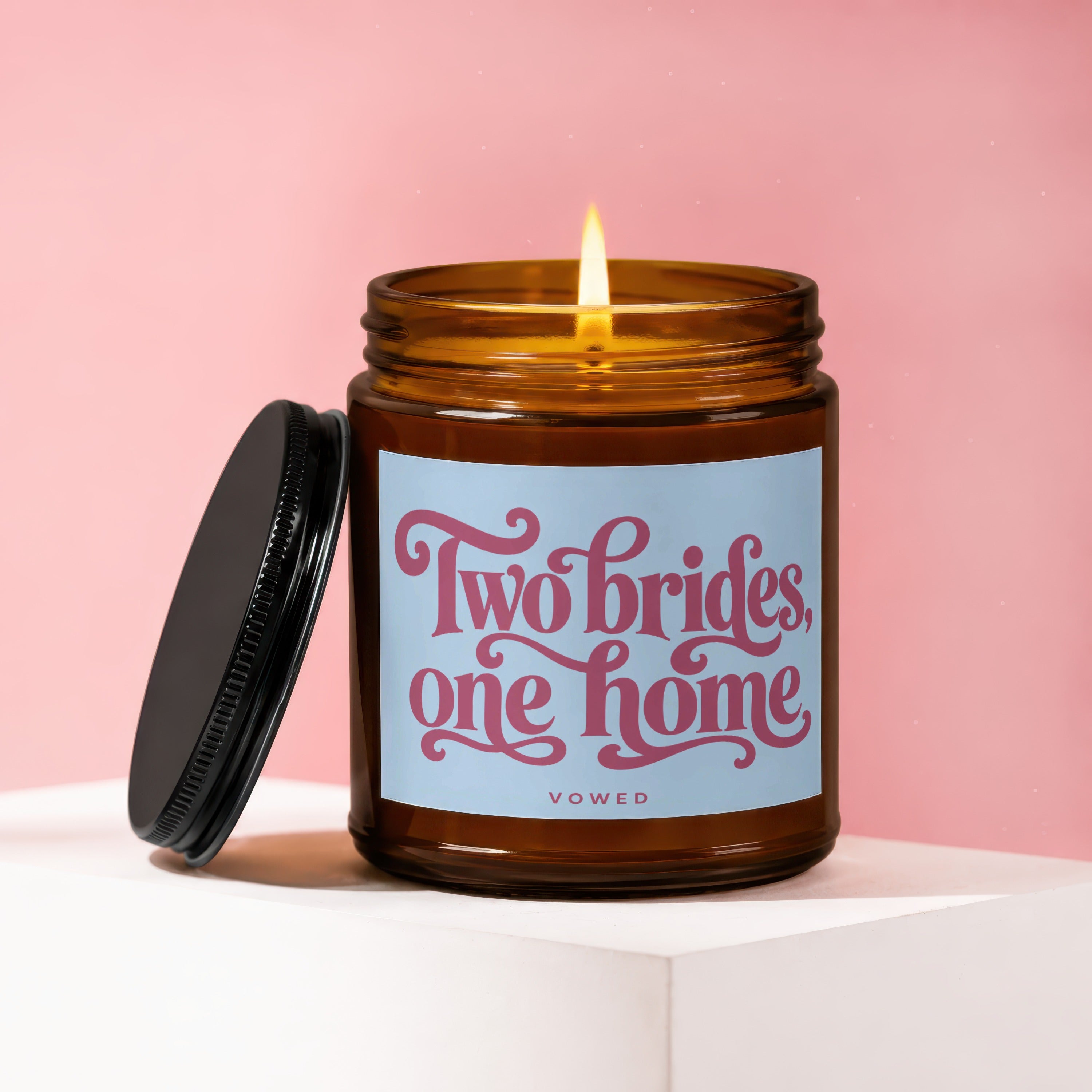 Amber jar candle with lit flame, black lid, and "Two brides, one home VOWED" text.