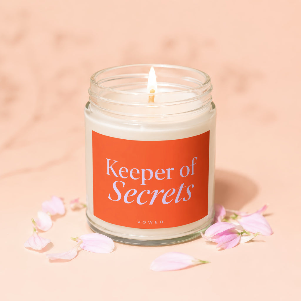 Glass jar candle with lit wick, orange label reading "Keeper of Secrets"