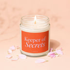 Glass jar candle with lit wick, orange label reading "Keeper of Secrets"