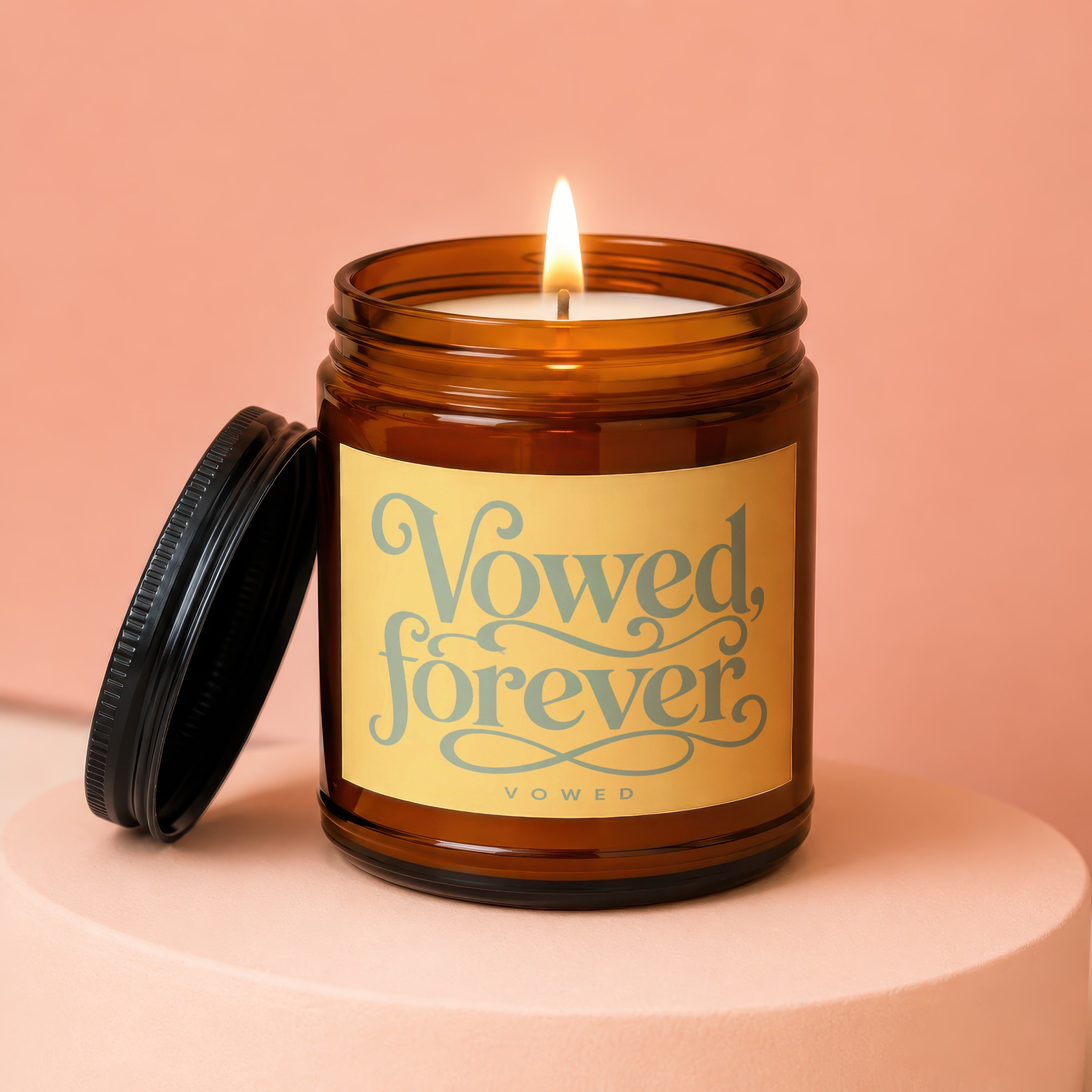 Lit amber glass candle with lit wick, label reads "Vowed, forever", lid off.