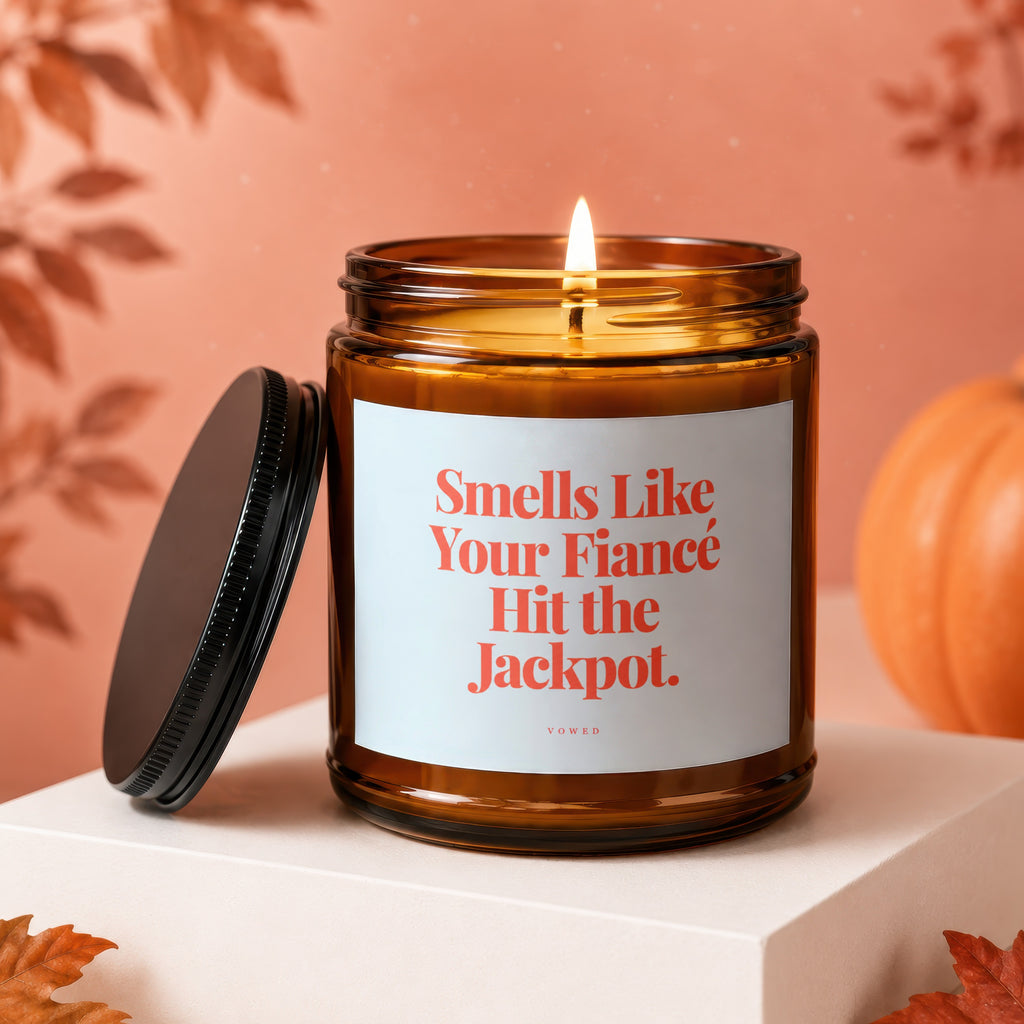 Amber candle with lit wick, reads "Smells Like Your Fiancé Hit the Jackpot."