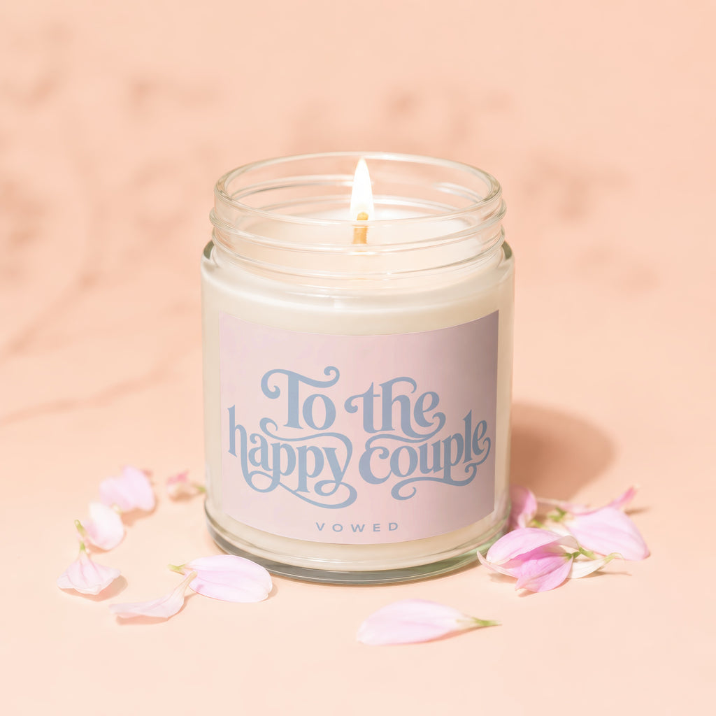 Lit candle with "To the happy couple vOWeD" text on label, surrounded by pink petals.