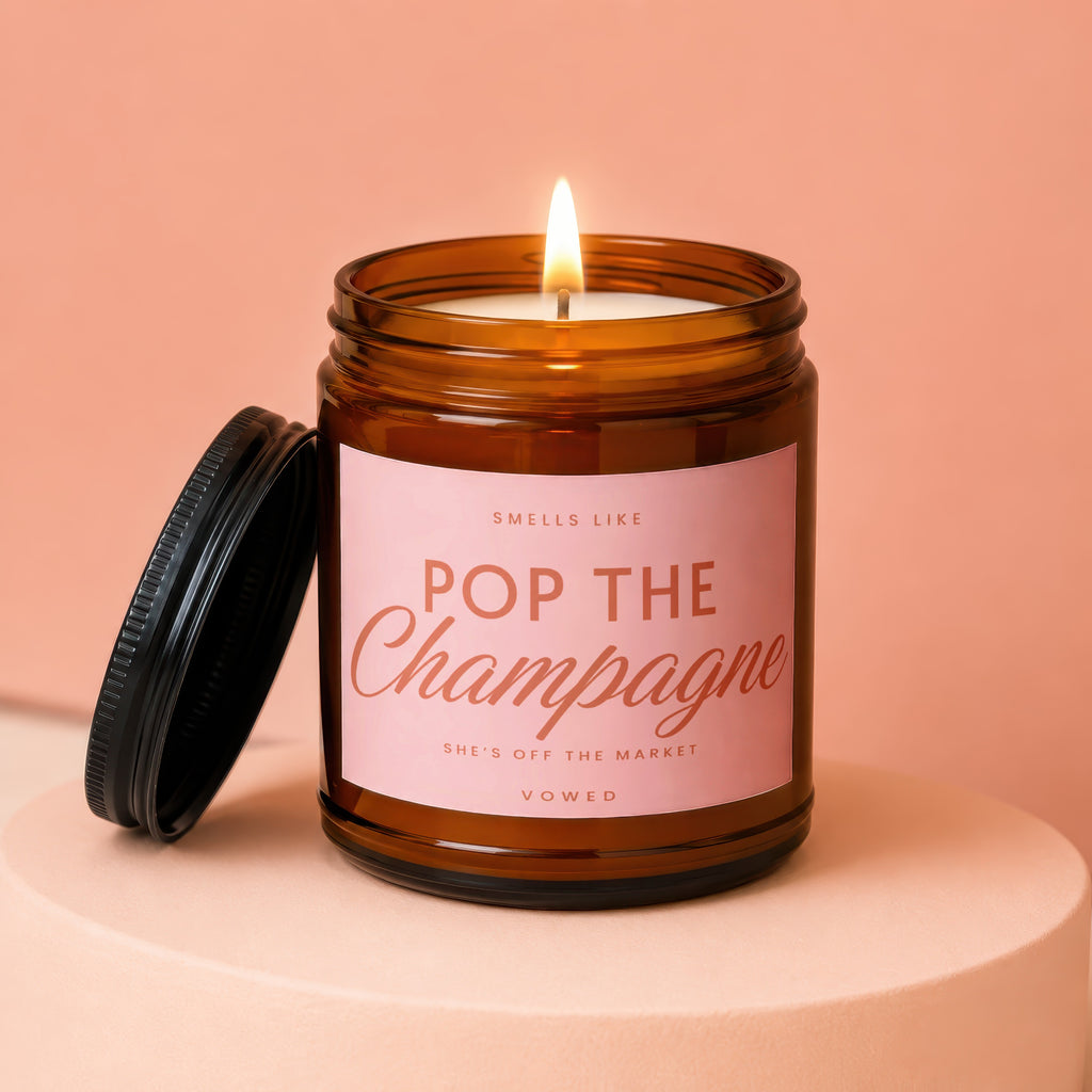 Amber jar candle with pink label that reads "Smells like Pop the Champagne She's off the market Vowed".