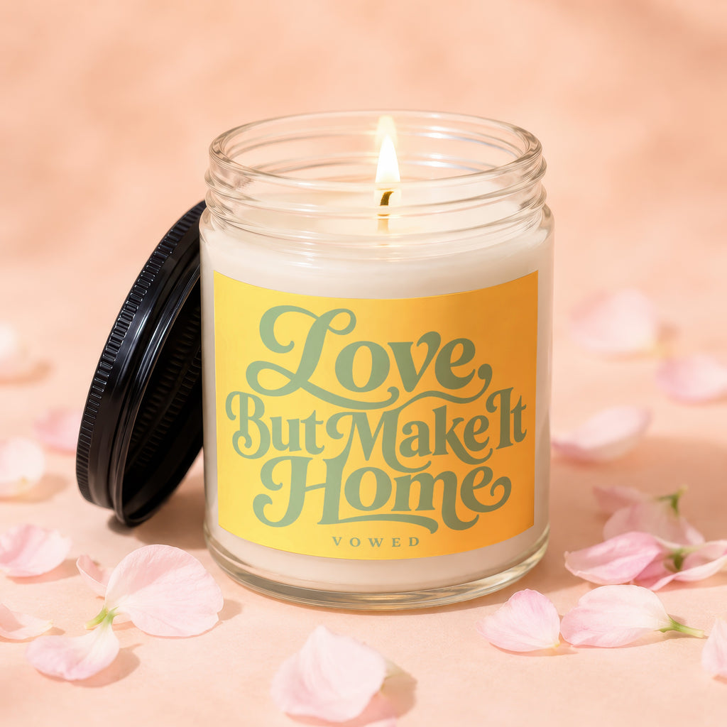 Glass candle with lit wick and yellow label reading "Love, But Make It Home".