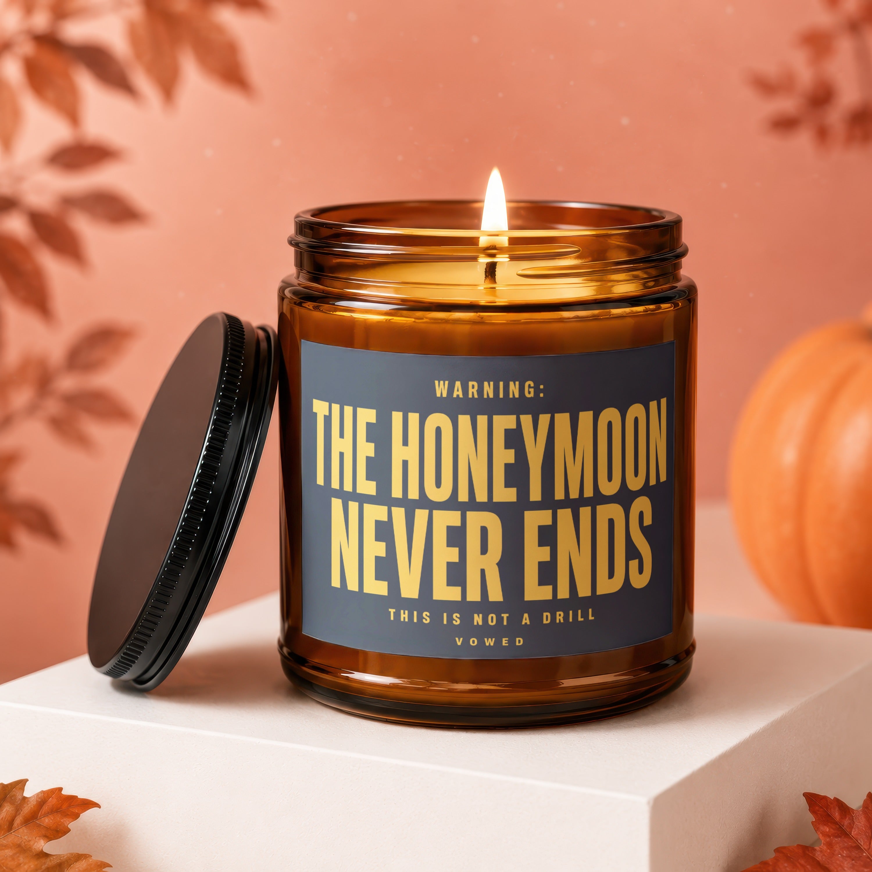 Amber jar candle with a lit wick and a black lid, reading "WARNING: THE HONEYMOON NEVER ENDS THIS IS NOT A DRILL VOWED".