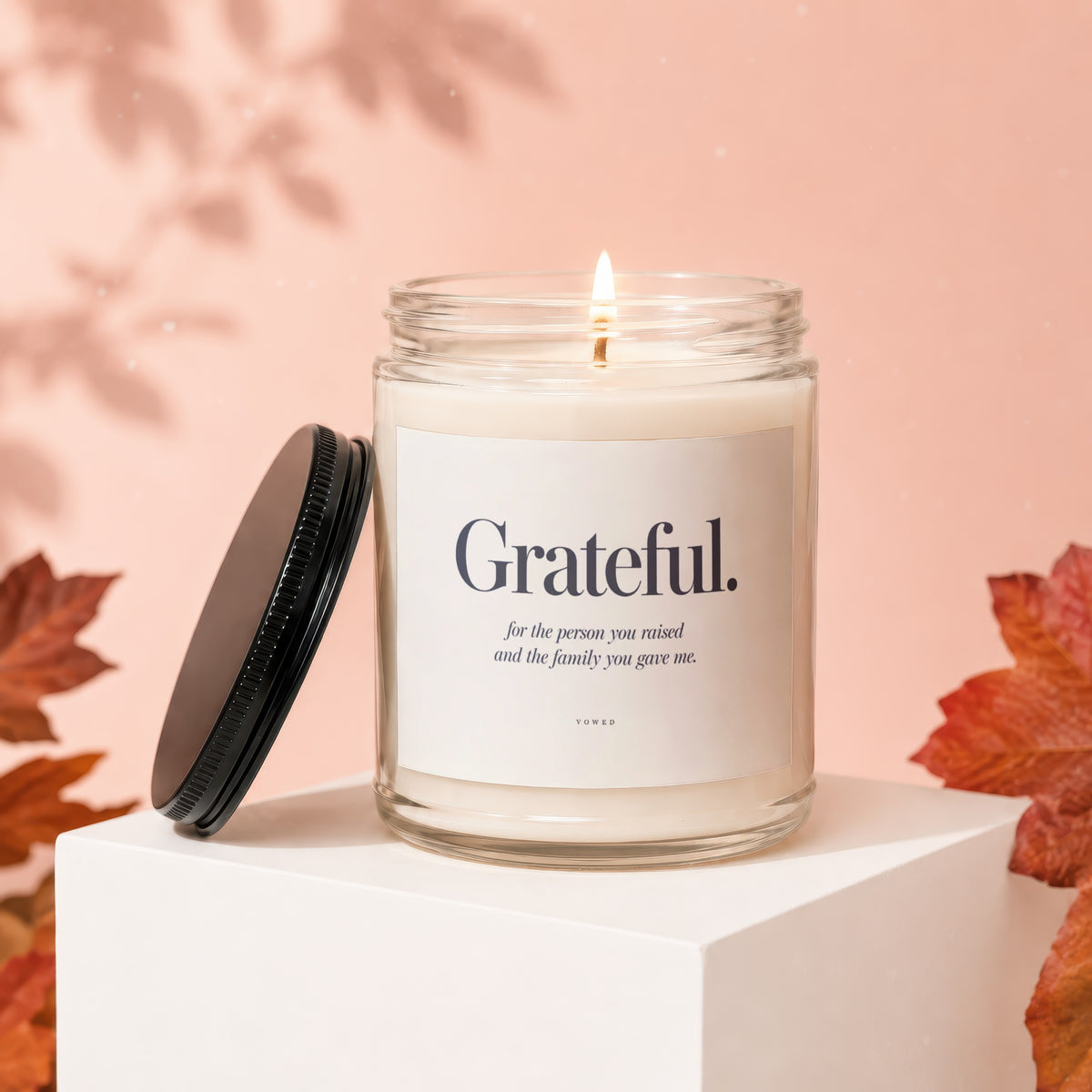 Candle with "Grateful" text on label, lit flame, black lid, and autumn leaves.