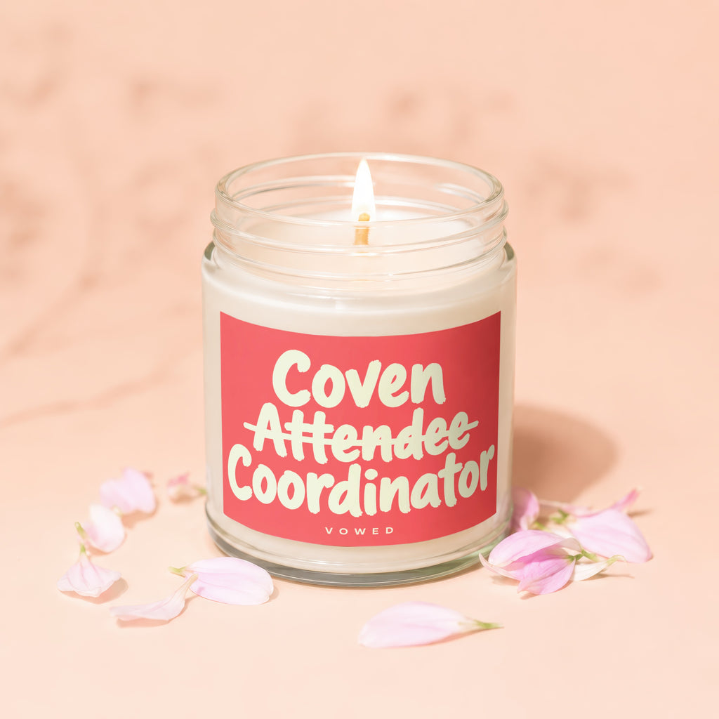 Candle with "Coven Attendee Coordinator" and "VOWED" text on pink label, lit flame.