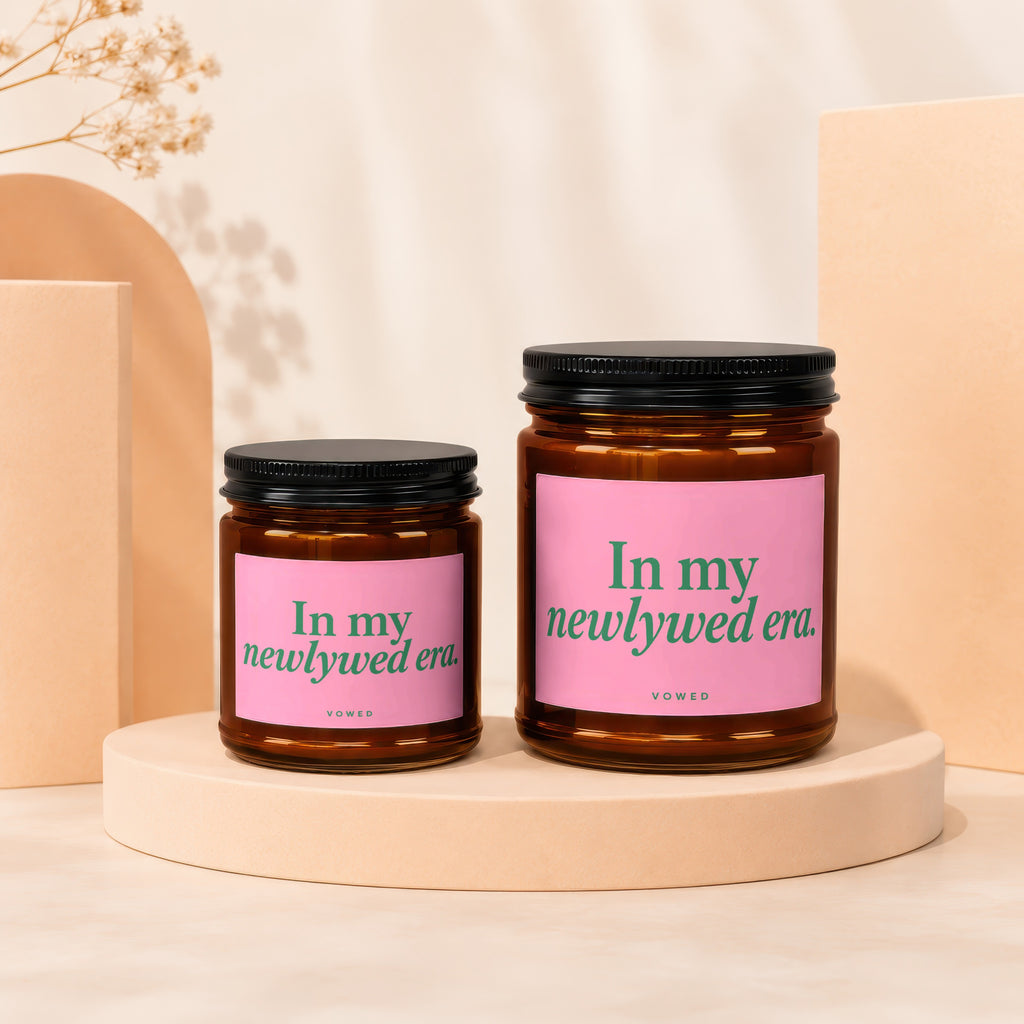Two amber glass candles with pink labels reading "In my newlywed era."
