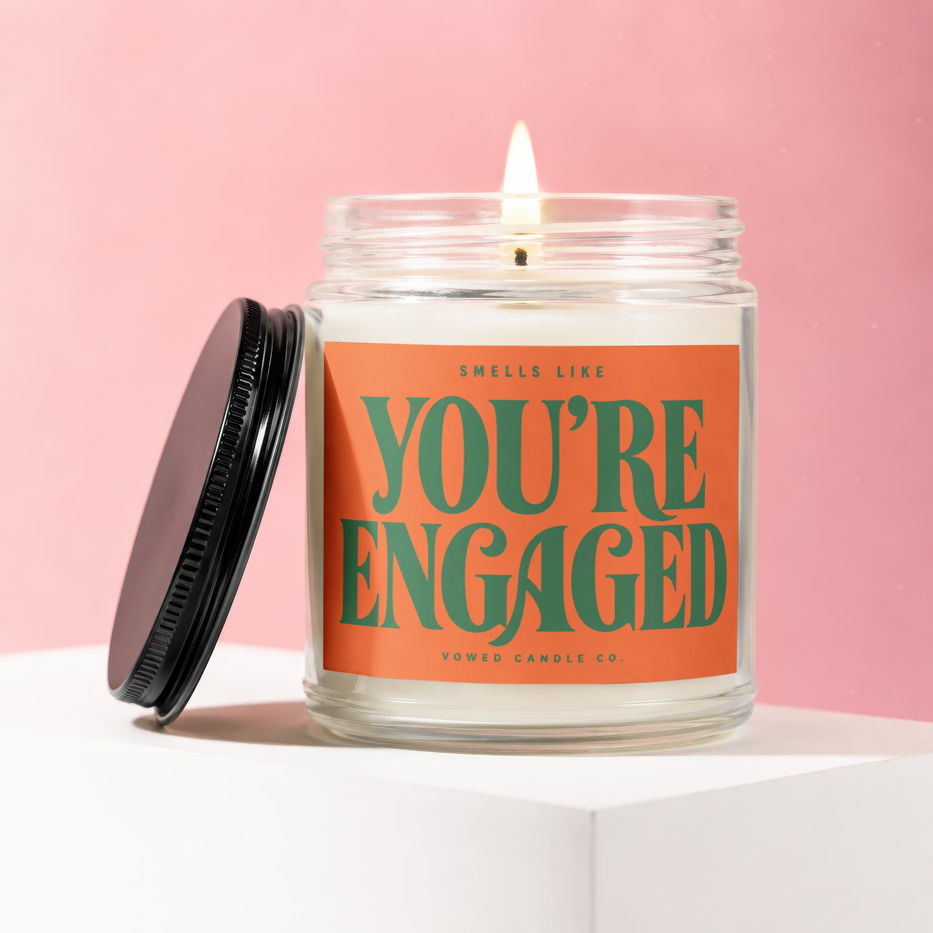 Jar candle with a lit wick and an orange label reading "Smells Like You're Engaged".
