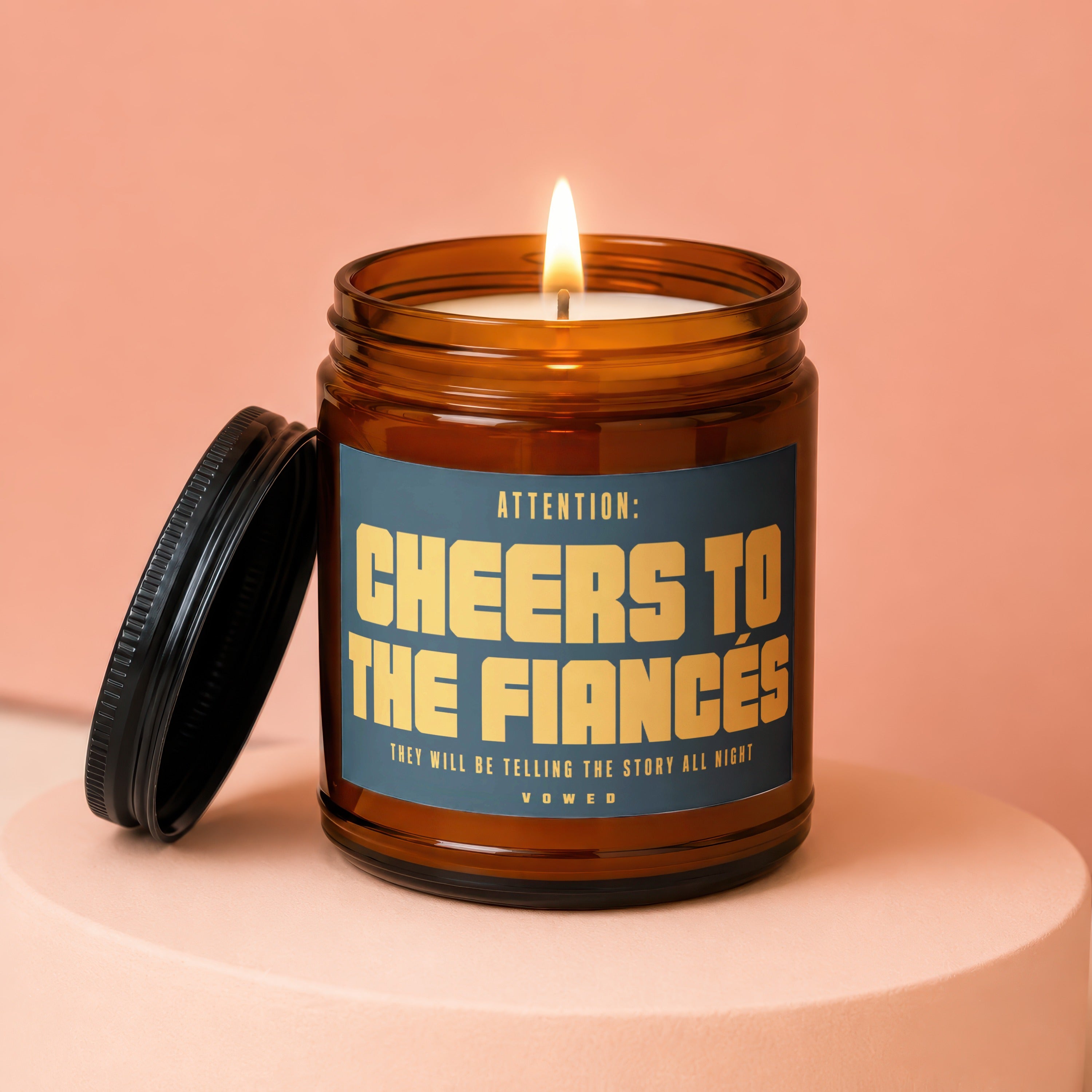 Amber candle with lit flame, black lid, and label reading "Cheers to the Fiances".