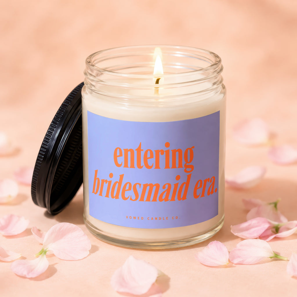 Lit candle with "entering bridesmaid era" text and "Vowed Candle Co." on a peach background with petals.