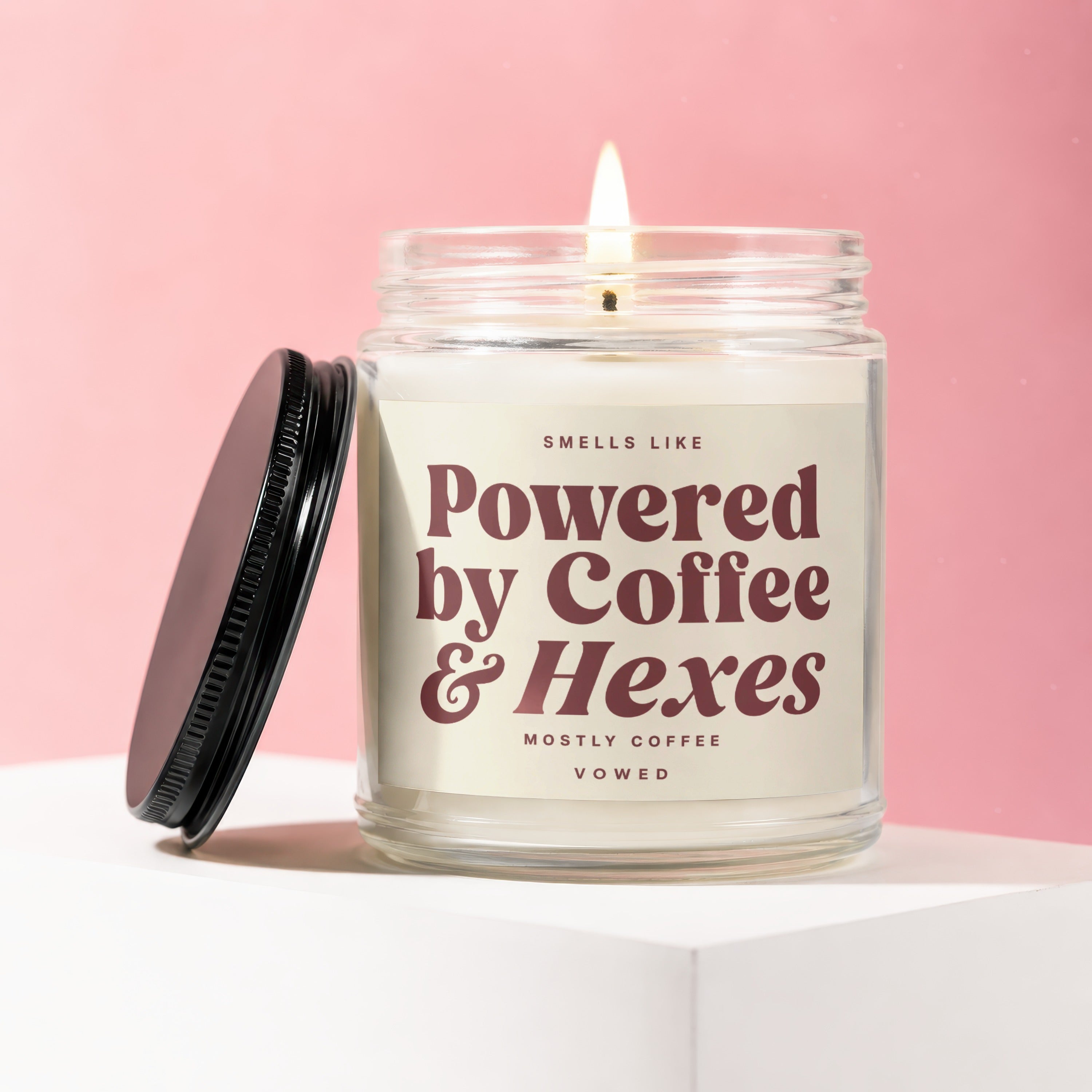 Lit candle in a glass jar with lid off, reading "Powered by Coffee & Hexes".