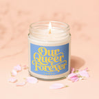 Jar candle with a blue label displaying "Our Queer Forever" in yellow text with pink petals scattered around.