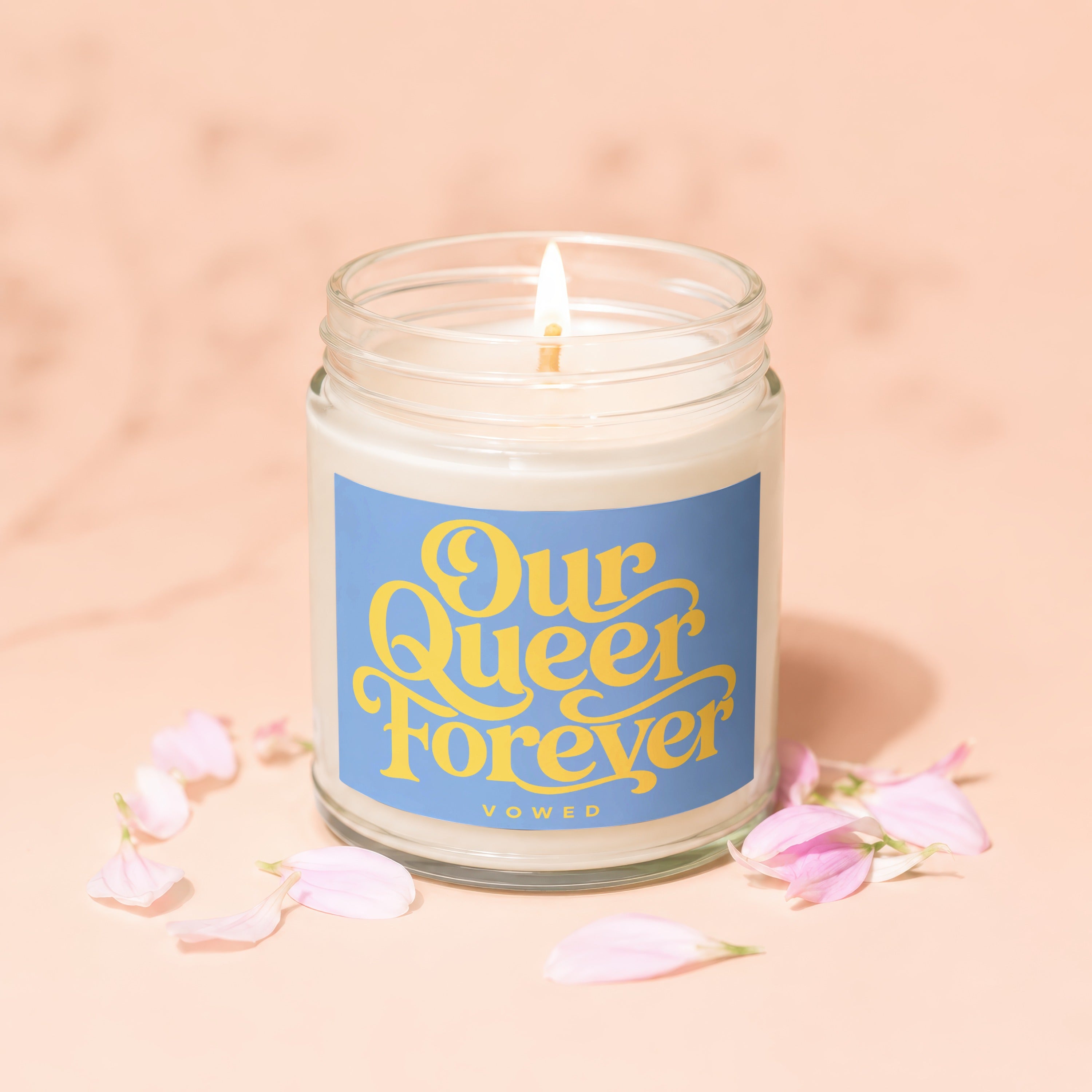 Jar candle with a blue label displaying "Our Queer Forever" in yellow text with pink petals scattered around.