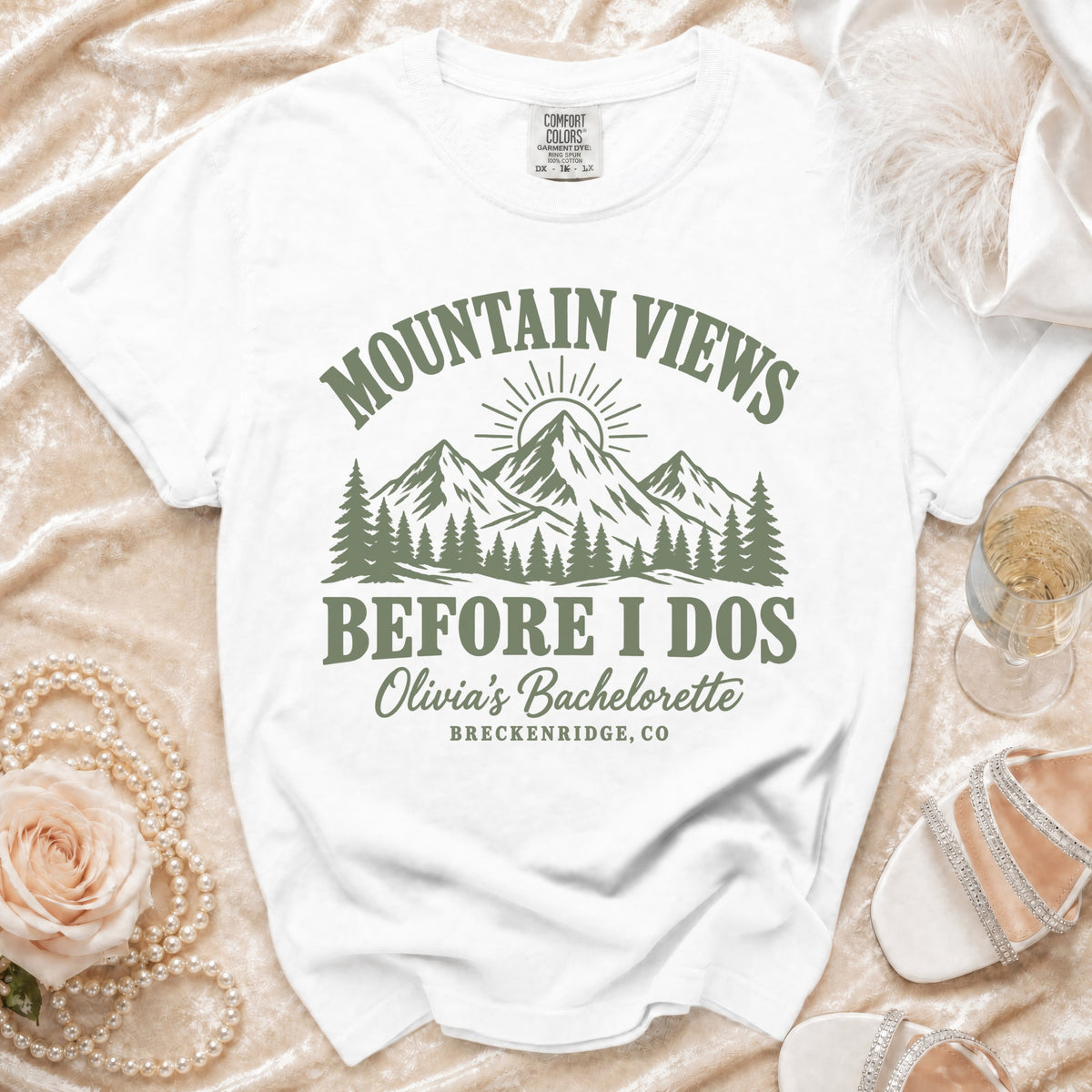 A white t-shirt featuring a green mountain landscape graphic and the text Mountain Views Before I Dos, Olivia's Bachelorette, Breckenridge, CO.