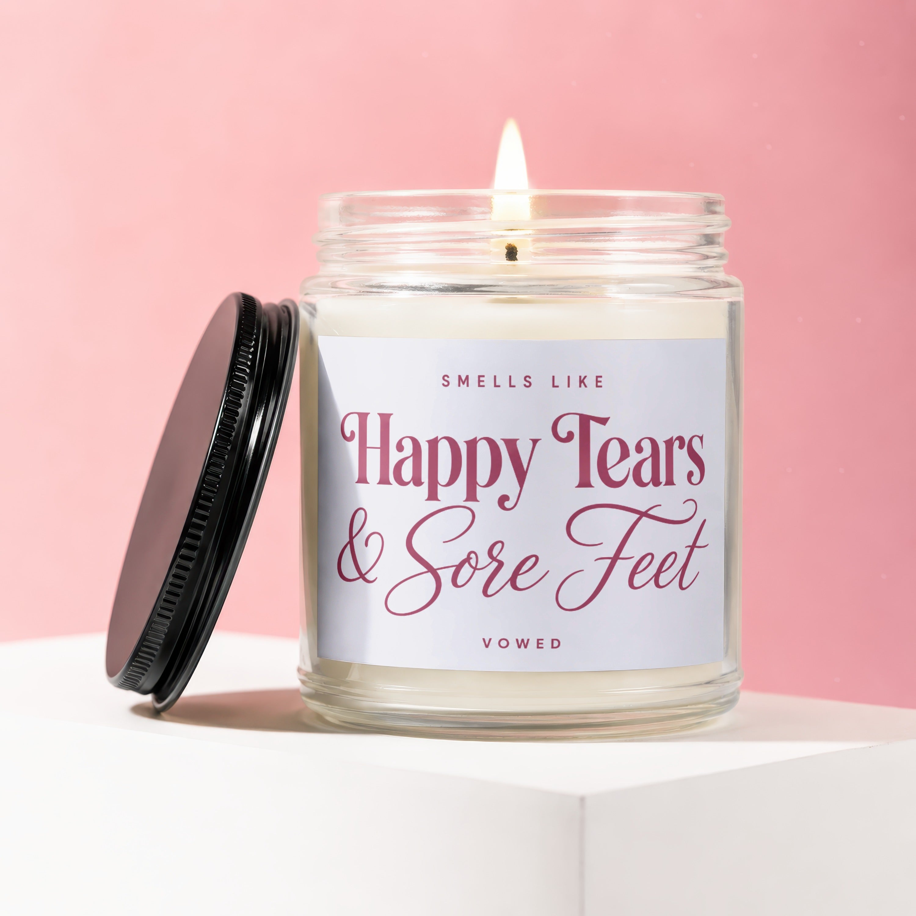 A lit candle in a glass jar, reading "Smells Like Happy Tears & Sore Feet".