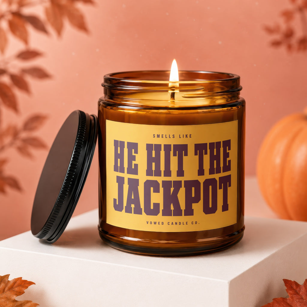 Amber glass candle with a lit wick and a yellow label reading "HE HIT THE JACKPOT".