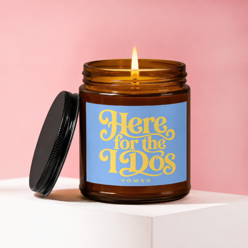 Amber jar candle with lit flame and blue label reading "Here for the Dos."