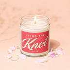 Lit candle in a glass jar with "TYING THE Knot" text and pink petals.