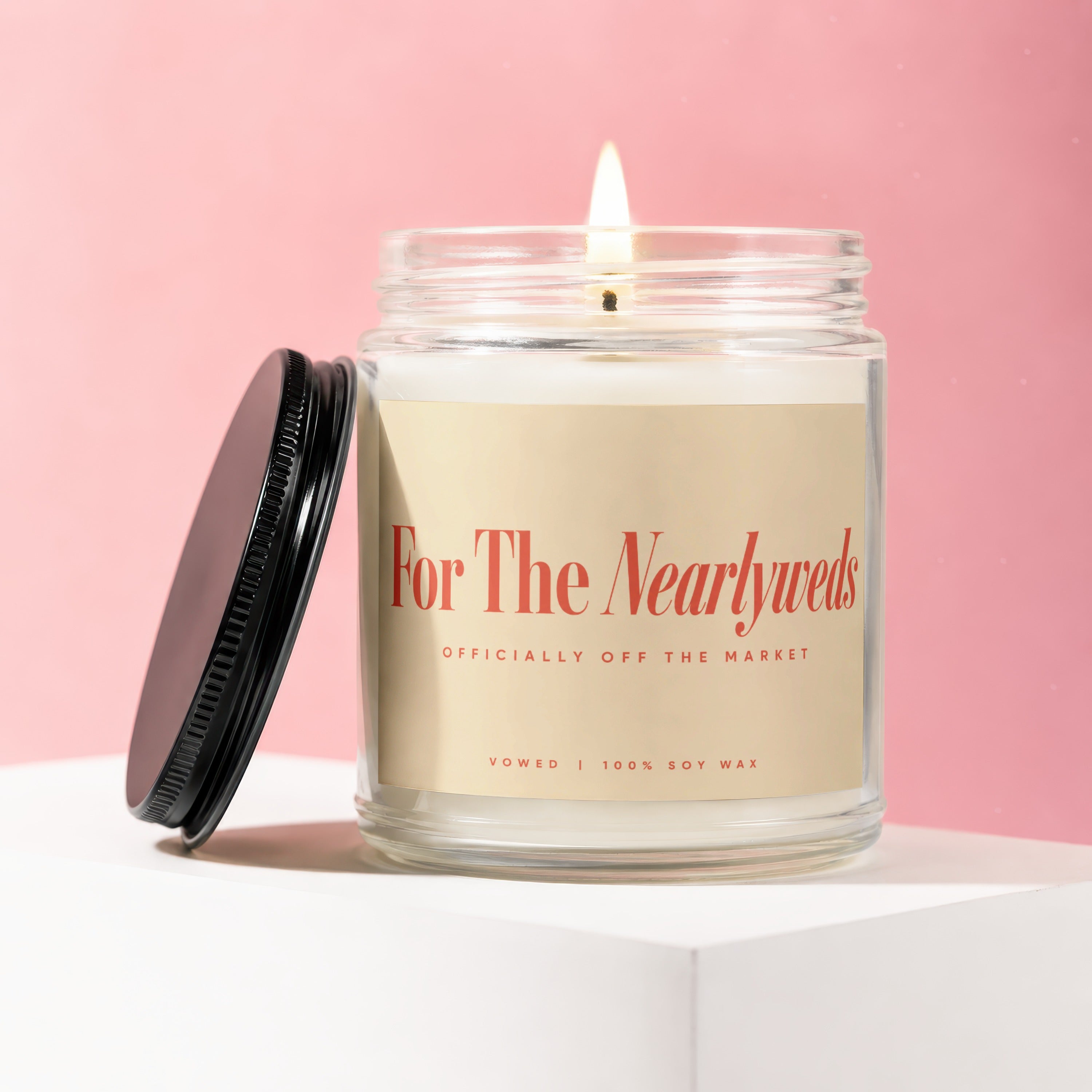 Lit candle in a glass jar labeled "For The Nearlyweds" on a white surface.