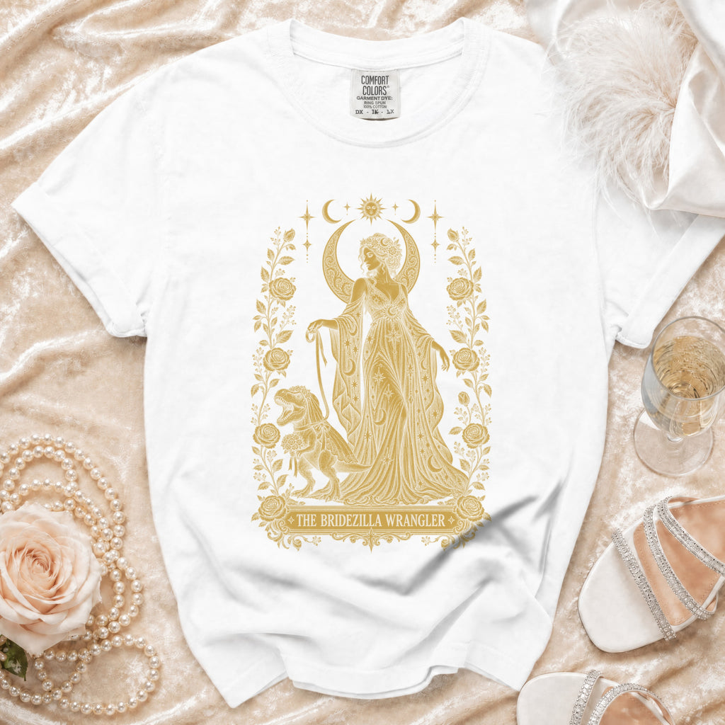 White t-shirt with gold print of a woman and a dinosaur. Text reads "The Bridezilla Wrangler".