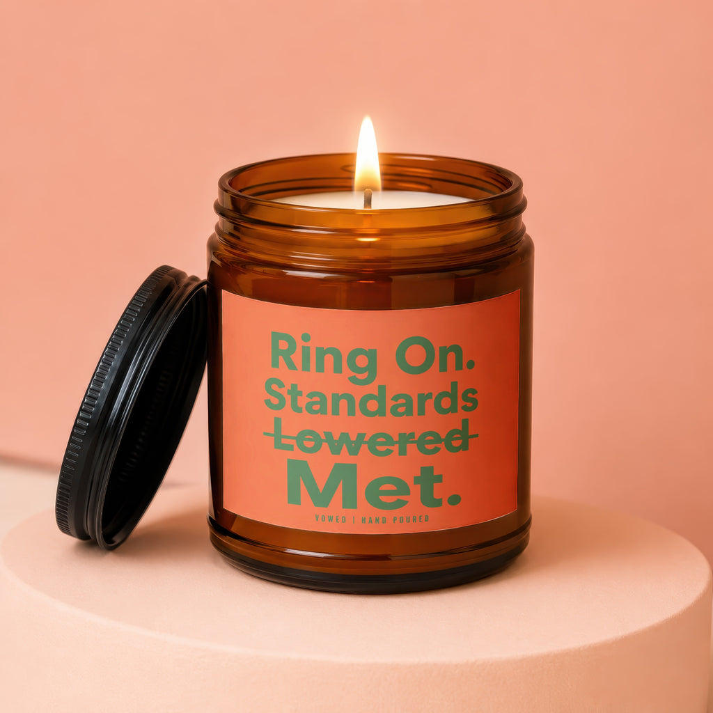 Amber glass candle with lit wick and black lid. Label reads "Ring On. Standards Lowered Met."