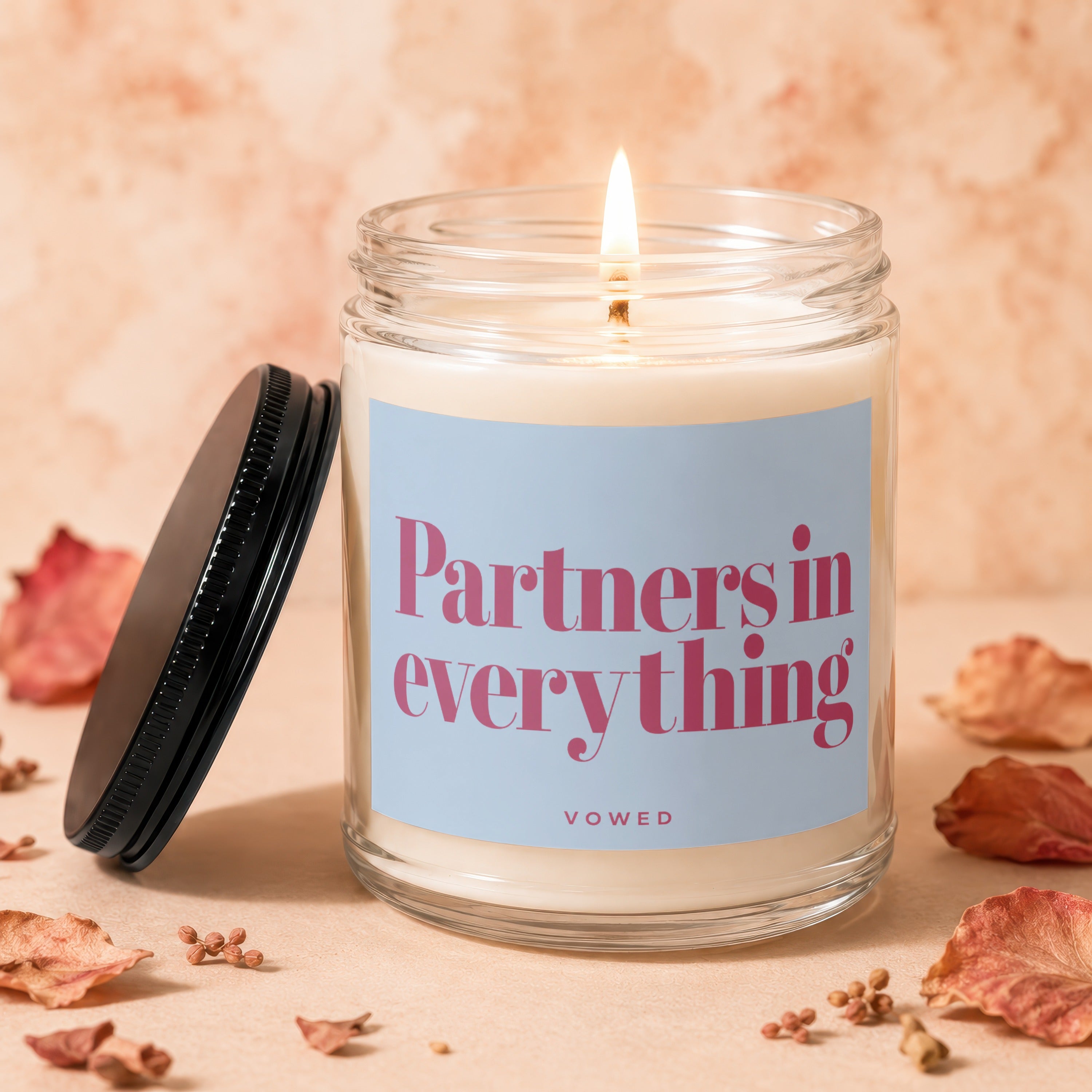 Glass candle with lit flame, blue label reads "Partners in everything" and "VOWED".