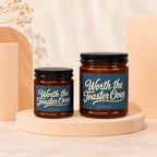 Two amber glass candles with black lids, blue labels reading "Worth the Toaster Oven."