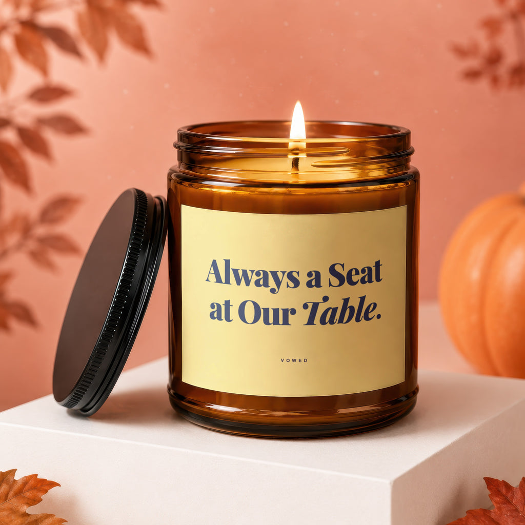Amber candle with lit wick, cream label reads "Always a Seat at Our Table. VOWED."