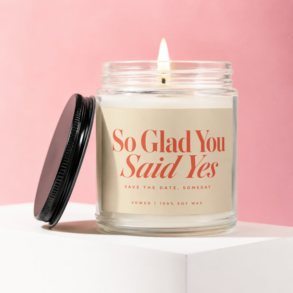 Glass jar candle with lit flame has "So Glad You Said Yes" text and 100% soy wax.