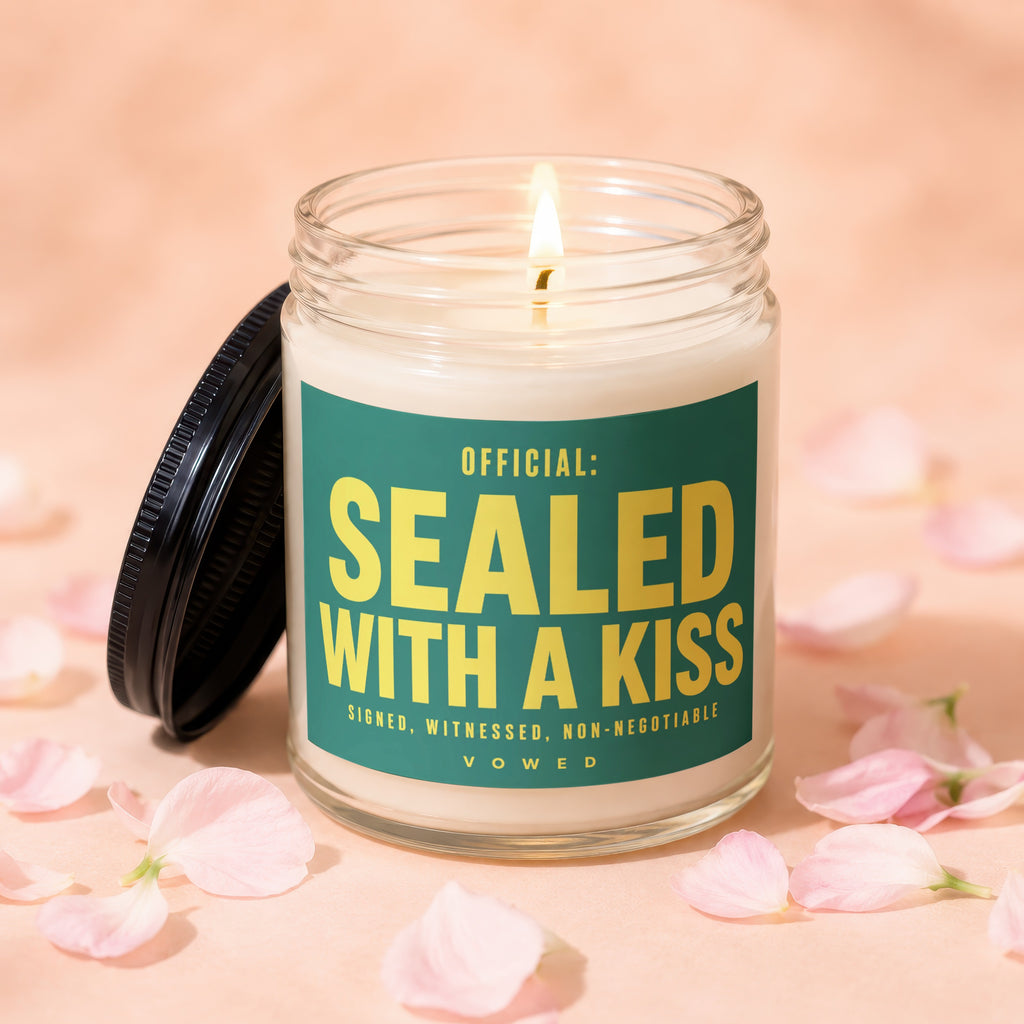 Glass candle with lit wick and label reading "OFFICIAL: SEALED WITH A KISS"