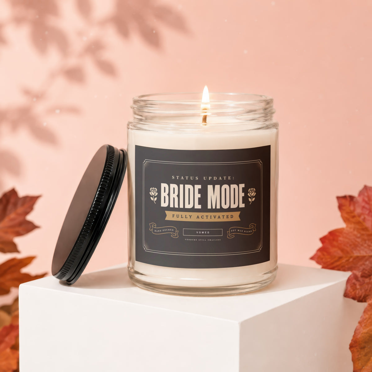 Glass candle with "Bride Mode Fully Activated" label, lid off, set among autumn leaves.