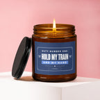 Amber jar candle with lit wick, black lid, and label text "HOLD MY TRAIN AND MY HAND".