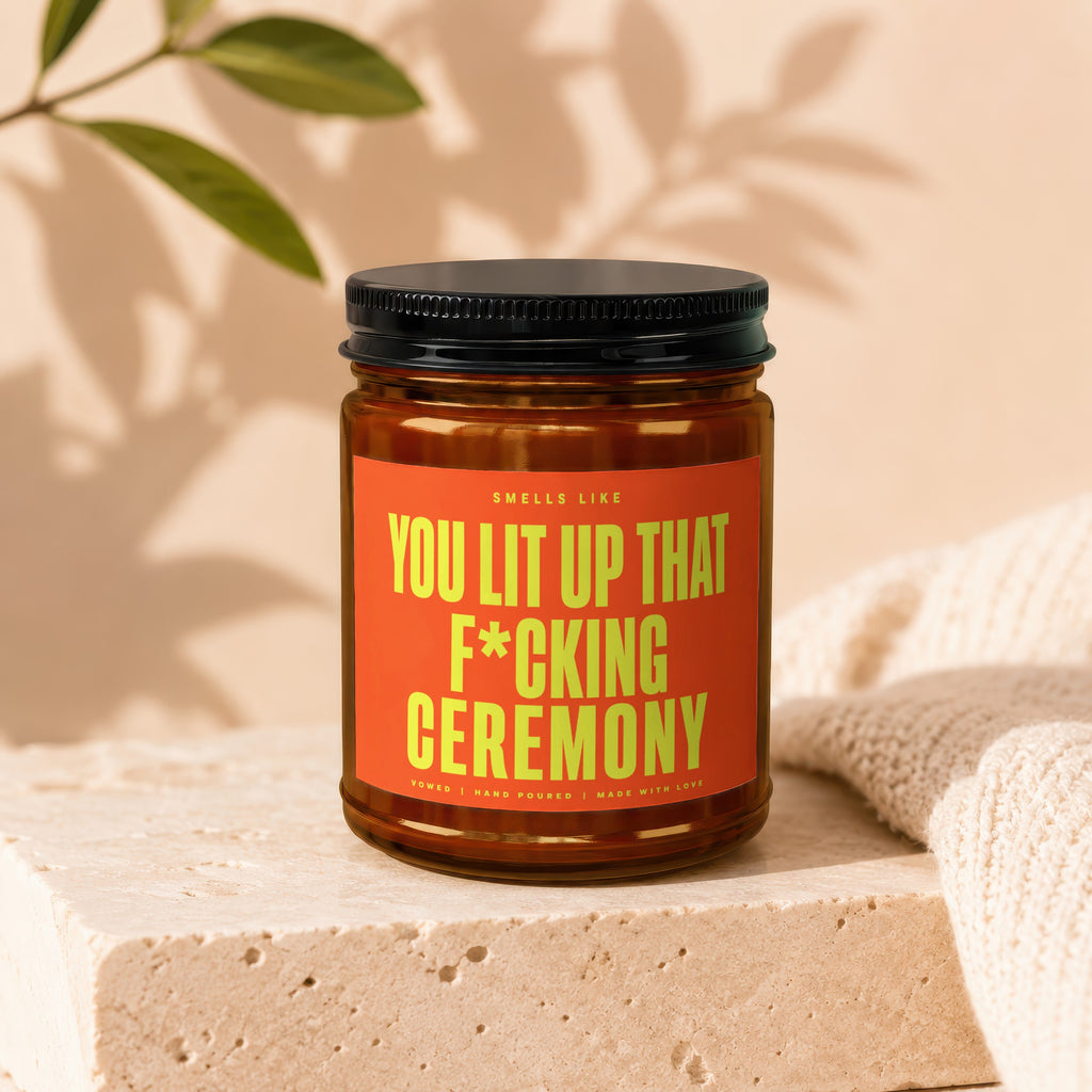 Amber glass candle with black lid and orange label reading "Smells Like You Lit Up That F*cking Ceremony".
