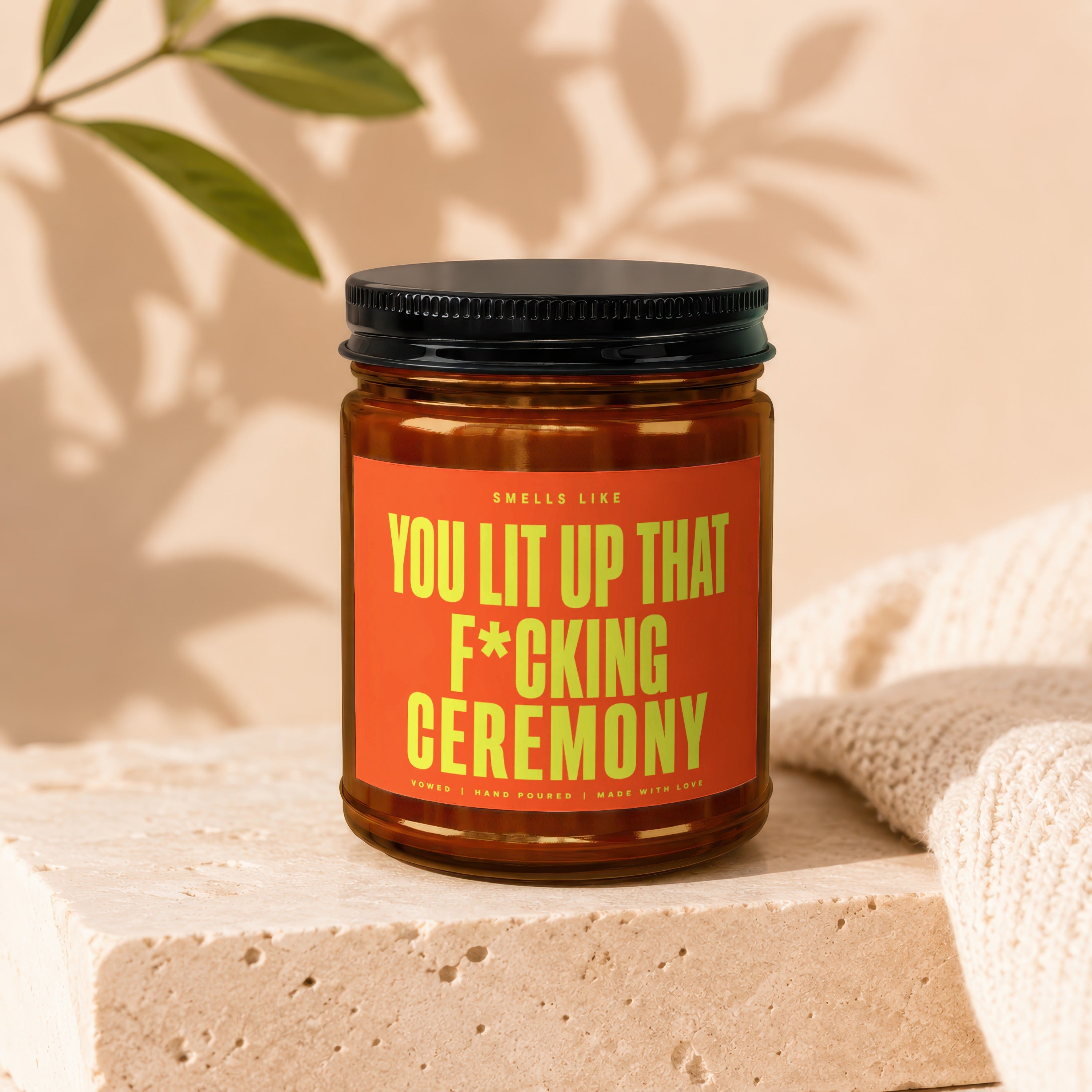Amber glass candle with black lid and orange label reading "Smells Like You Lit Up That F*cking Ceremony".
