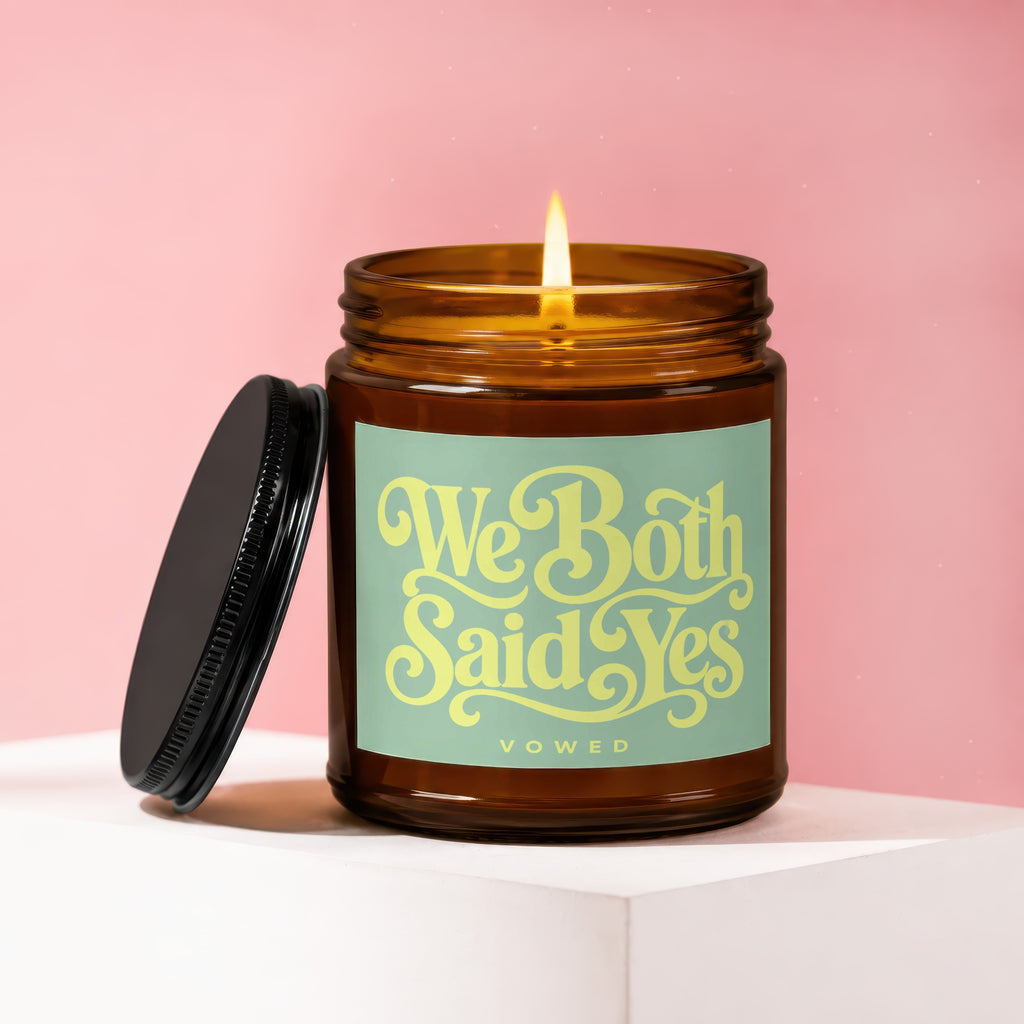 Amber glass candle with lit flame and lid beside it, reads "We Both Said Yes VOWED".