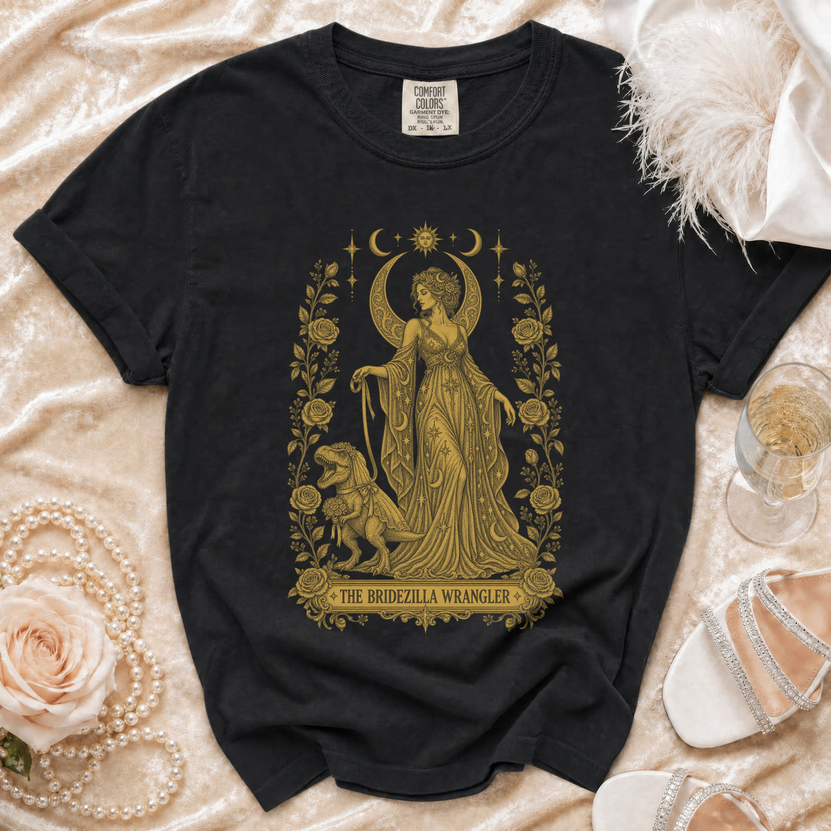 Black t-shirt with golden graphic of a woman and dinosaur, "The Bridezilla Wrangler" text.
