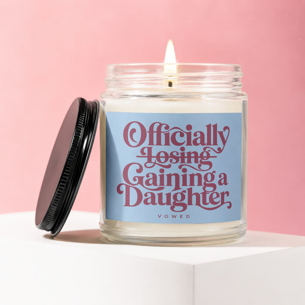 Glass candle with "Officially Losing, Gaining a Daughter" text and a lit wick.