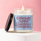 Glass candle with "Officially Losing, Gaining a Daughter" text and a lit wick.