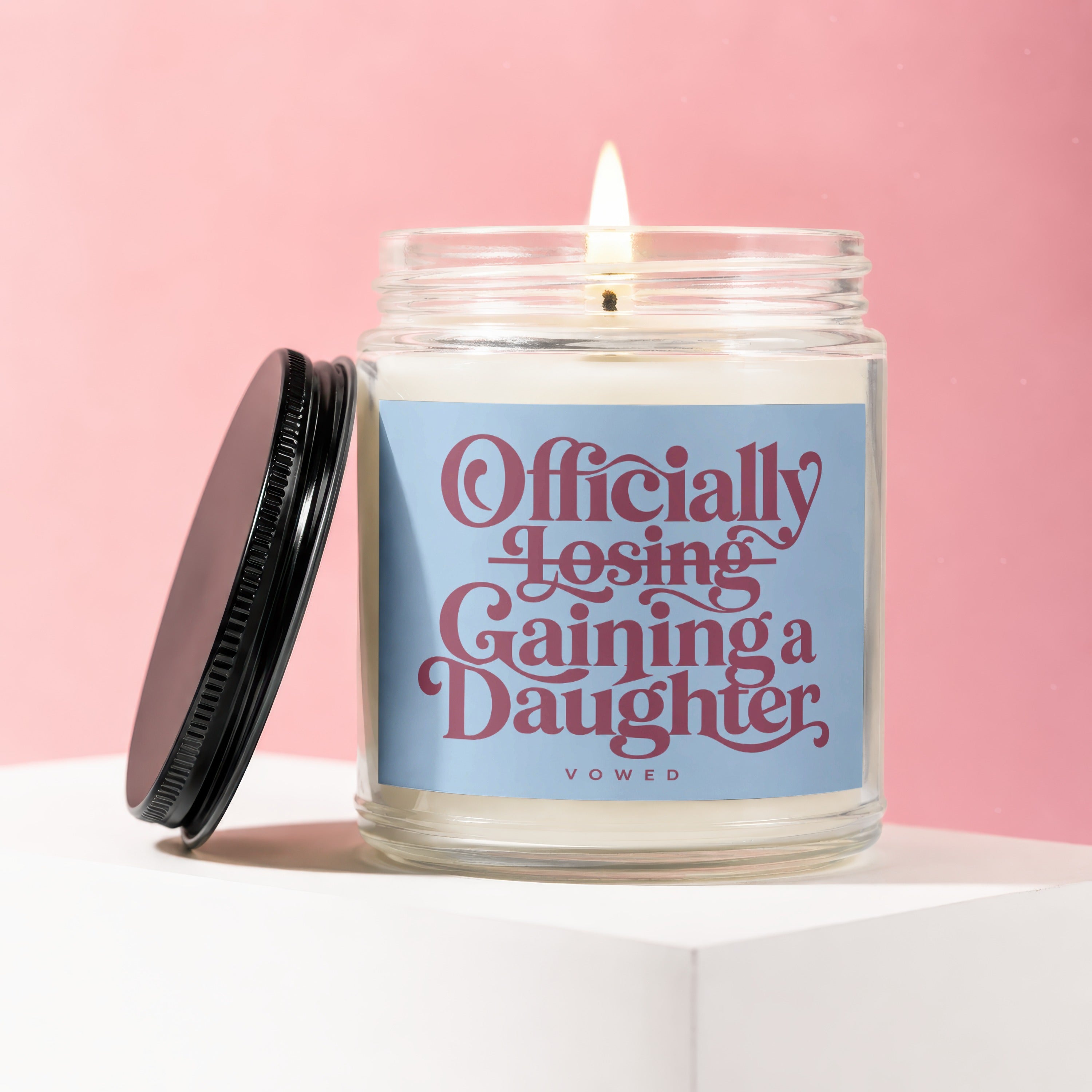 Glass candle with "Officially Losing, Gaining a Daughter" text and a lit wick.