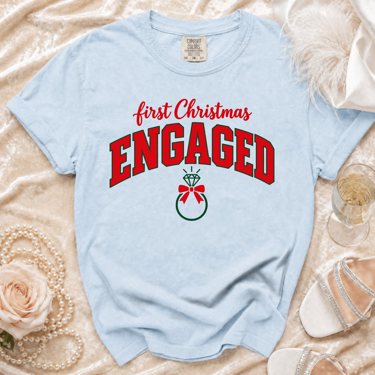 Light blue t-shirt featuring red and green text reading First Christmas Engaged, surrounded by decorative accessories.