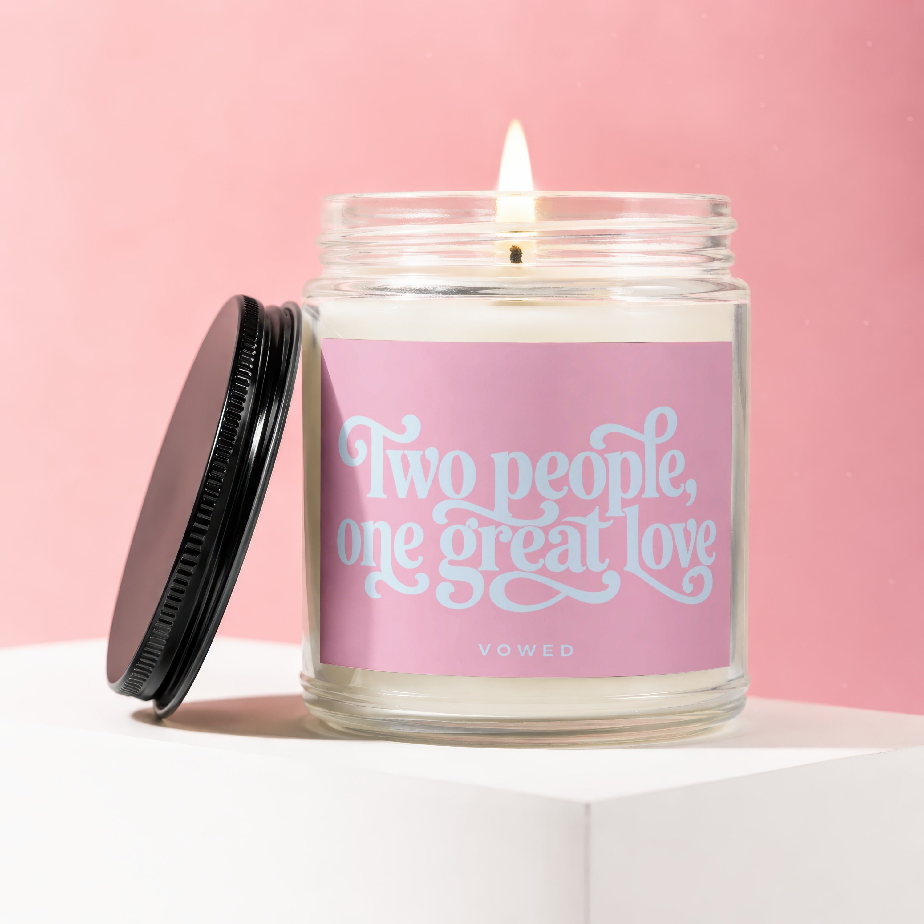 Glass jar candle with a lit wick, featuring a pink label with "Two people, one great love" in light blue script.