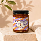 Amber candle with black lid, featuring a wavy orange and purple label that reads "CHAMPAGNE ON ME! WHEN YOU SAY YES! VOWED".
