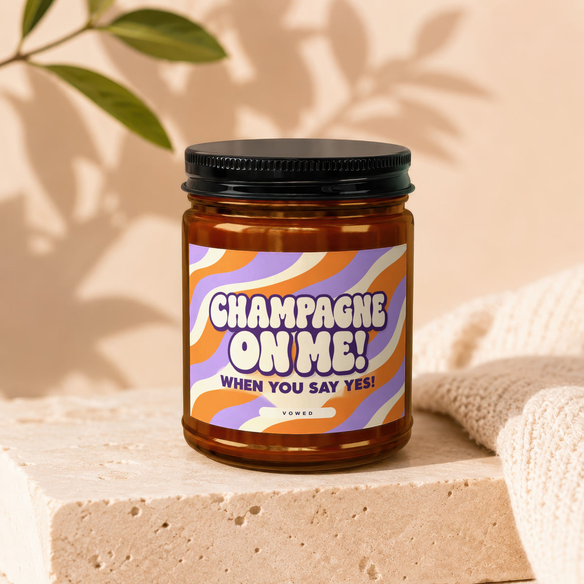 Amber candle with black lid, featuring a wavy orange and purple label that reads "CHAMPAGNE ON ME! WHEN YOU SAY YES! VOWED".