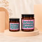 Two amber jar candles with pink labels saying "Surviving Celebrating Forever" and "VOWED".