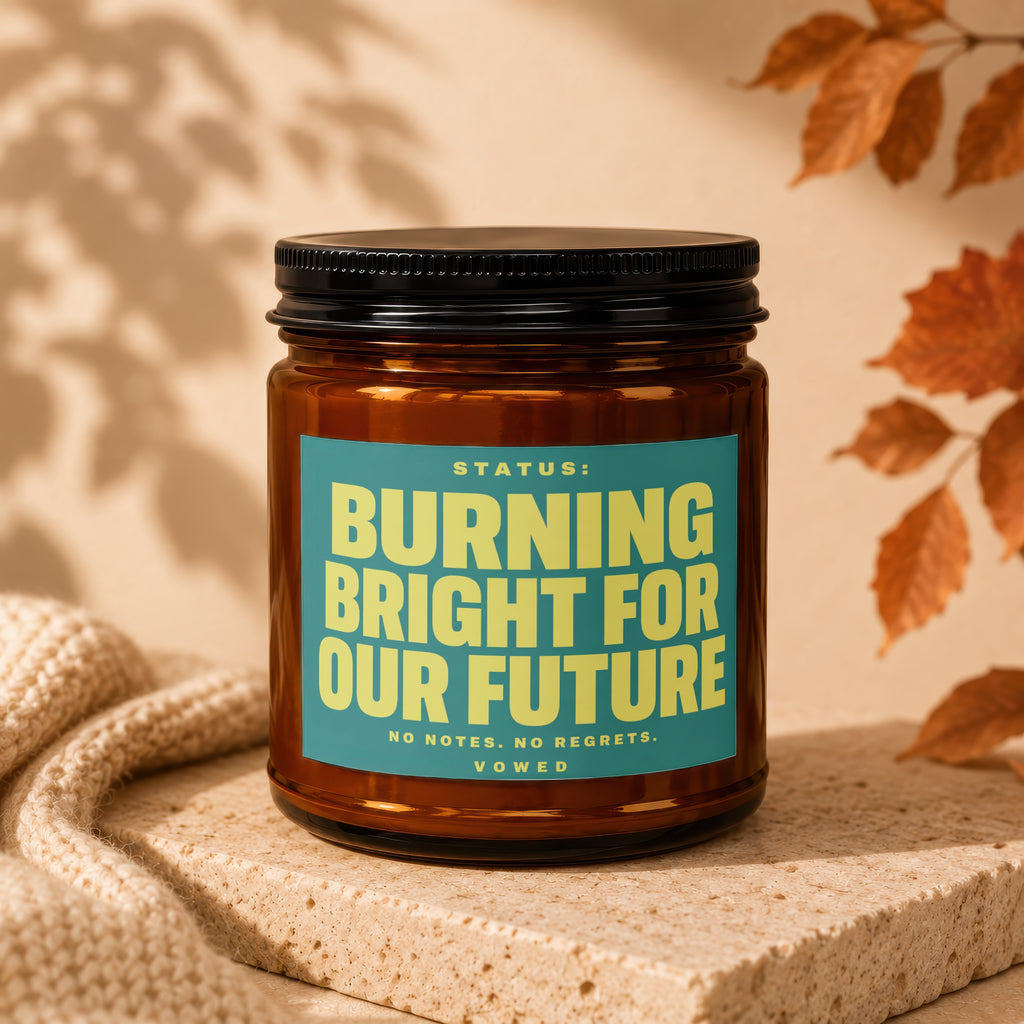Amber jar candle with teal label reading "STATUS: BURNING BRIGHT FOR OUR FUTURE".